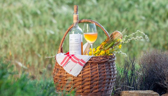 A picnic basket in a field with a transparent bottle of a white wine, a glass with some intense yellow coloured wine, some bread and flowers.
