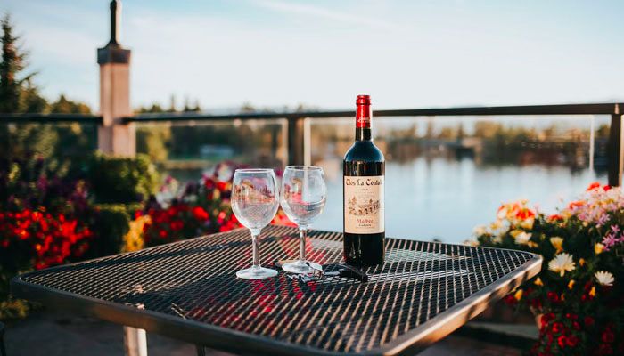 A bottle with a classic red wine label and two empty glasses on a table with river view on a terrasse.