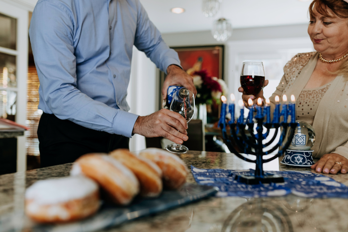 Man pouring himself a glass of red wine and woman holding a glass full of red wine, during the Jewish celebration of Hanukkah. On the table, a tray with sweets and a hanukkiah (9-branched candelabra) with nine lit blue candles. Photo by Rodnae Productions from Pexels.