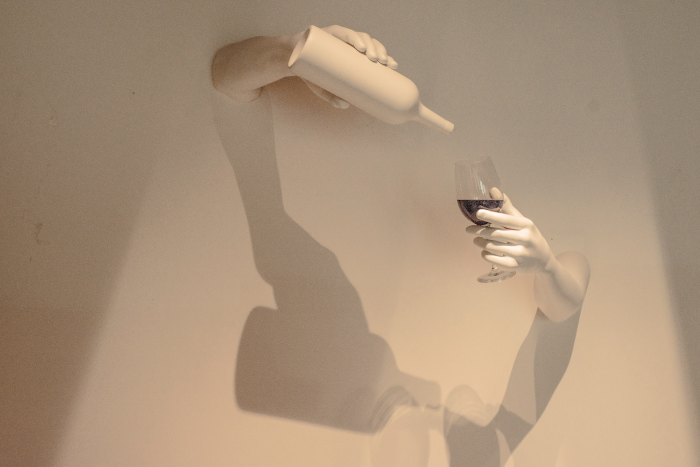 Artwork, hands coming out of two holes in a wall, pouring red wine into a glass from a completely white bottle. Photo by Hayffield L. from Unsplash