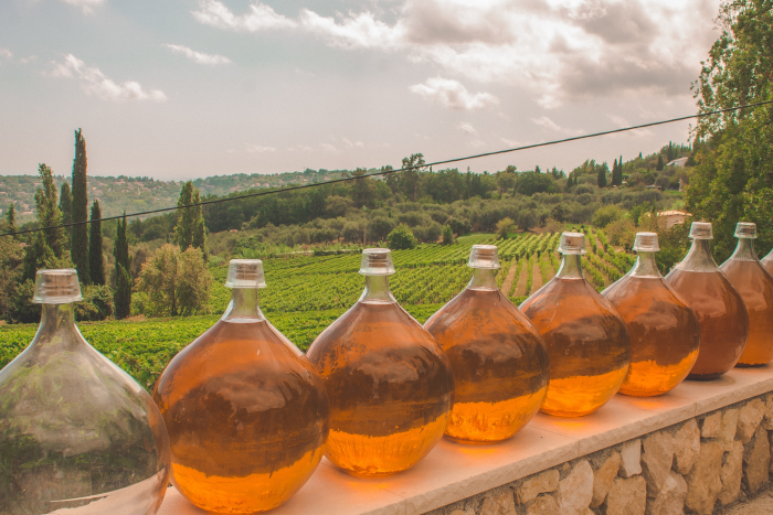 Rounded clear glass carafes filled with orange wine, resting on a stone wall. In the background, clusters of trees enveloping the vineyards. Greyish blue sky with threatening storm clouds. Photo by Diane Picchiottino from Unsplash.