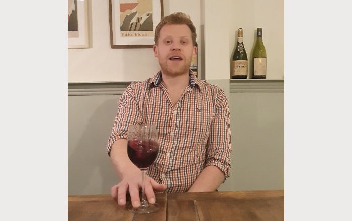 Sommelier holding a glass of red wine with two fingers of his right hand and drawing circles to swirl the wine inside. In the background, two paintings on the wall and 2 bottles of wine on the shelf.