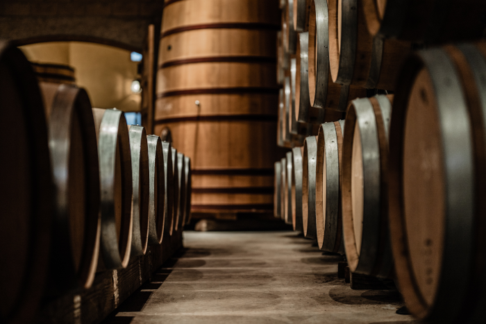 Rows of wine barrels in a cellar. In the background a large wooden tub. Photo by David Goldman from Unsplash.