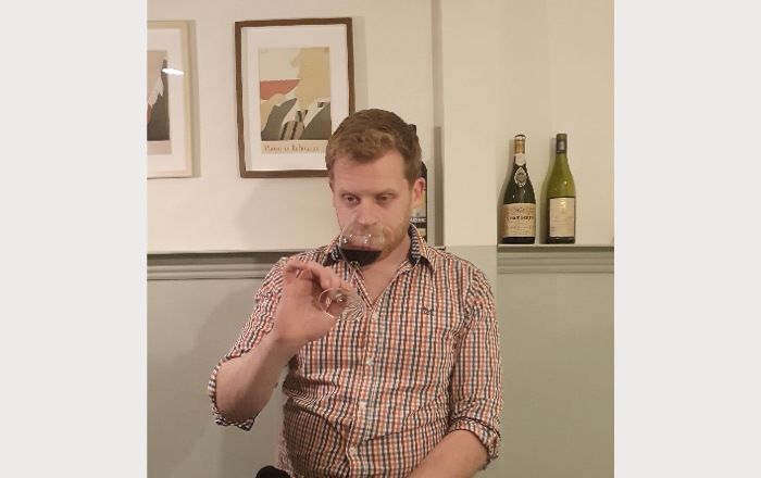 Sommelier sitting at a table, holding a glass of red wine between the fingers of his right hand and smelling the wine. In the background, two paintings on the wall and 2 bottles of wine on the shelf.