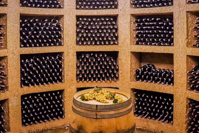 Circular wall full of niches filled with wine bottles for ageing. In the centre of the photo, standing upright, an old barrel with bunches of white grapes on top. Photo by Thomas George from Unsplash.