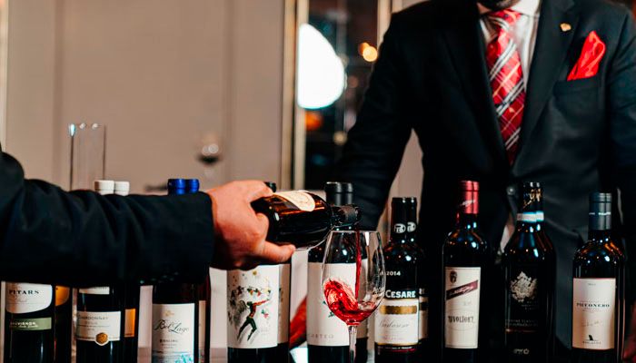 A bartender serving red wine to a man in front of a wide range of red wine bottles.