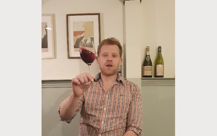 Sommelier holding a glass of red wine raised with his right hand and swirling the wine inside. In the background, two paintings on the wall and 2 bottles of wine on the shelf.