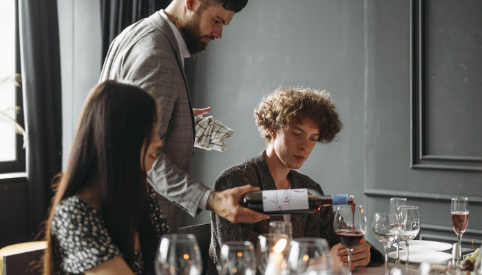 A man serving red wine into a glass to one of the guests at a table.