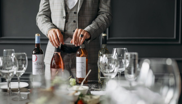 A person holding wine bottles on a laid table with empty wines glasses.