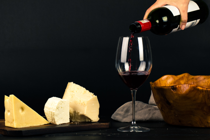 Photo of person pouring wine into glass besides some cheese pairings. Photo by Ray Piedra from Pexels.